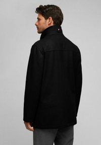 Schwarze Wollmischung Jacke mit hohem Kragen, geradem Schnitt und einer dezenten rückseitigen Naht. Verfügt über ein kleines farbiges Logo nahe des Kragens.