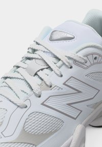 Κοντινό πλάνο ενός λευκού αθλητικού παπουτσιού με υφασμένο πλέγμα, χοντρές κορδόνια και μεγάλο λογότυπο "N" στο πλάι.