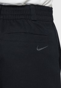 Čierne športové nohavice s elastickým pásom, majúce zadné vrecko a malé strieborné vyšívané logo Nike blízko pásu.