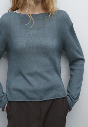 Person wearing teal knit sweater with long sleeves and dark pants, hands tucked into pockets, standing against a light-colored wall.