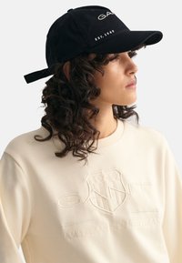 Black baseball cap features embroidered "GANT" logo and "EST. 1949" detail. Paired with a cream sweatshirt showcasing embossed brand emblem.