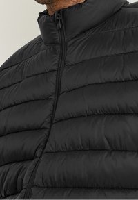 Czarna puchowa kurtka z wysokim kołnierzem, z poziomymi pikowanymi liniami, zamkiem błyskawicznym z przodu i gładką fakturą. Zaprojektowana z myślą o ciepłe i wygodzie.