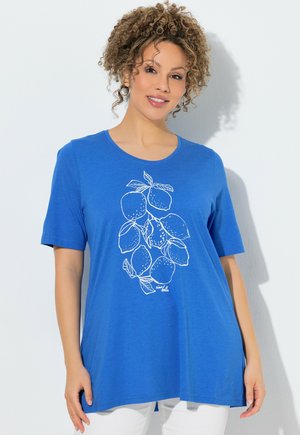 Kobieta o kręconych włosach, ubrana w niebieską koszulkę z białym wzorem gałązki cytryny, stojąca na jednolitym białym tle.