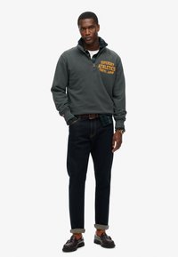 Hombre con sudadera de media cremallera en verde oscuro con el texto "Superdry Athletics Tokyo", combinada con una camisa de cuadros, jeans oscuros y mocasines marrones.