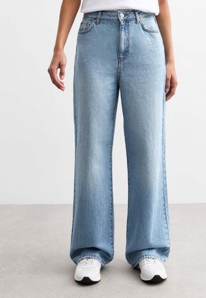 Person iført lyseblå vidde ben jeans, hvide sneakers og en hvid top, stående på en grå gulv mod en ensfarvet baggrund.