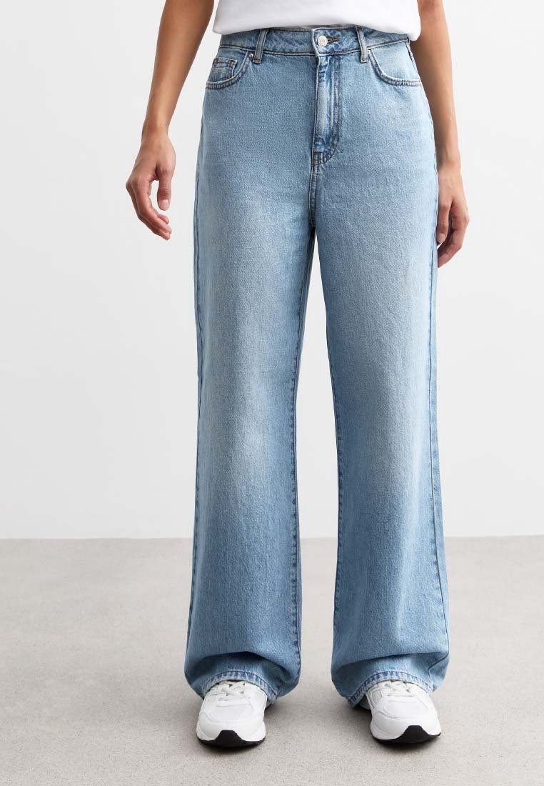 Personne portant un jean large bleu clair, des baskets blanches et un haut blanc, debout sur un sol gris devant un fond uni.