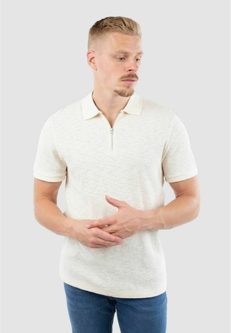 Kurzarm-Polo mit Reißverschlusskragen aus strukturiertem Creme-Stoff und beigem Kragen. Getragen mit blauen Jeans, die einen lässigen Schnitt und subtile Details aufweisen.