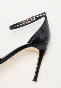 Zapato de tacón alto de charol negro con punta afilada y tira en el tobillo. Presenta un acento dorado con el logo en la tira. Textura suave.