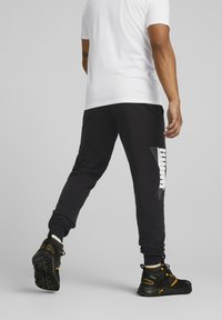 Pantalones deportivos negros con diseño ajustado y estampado de logo en el lateral, combinados con zapatillas deportivas negras y amarillas, llevados con una camiseta blanca.