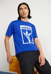 Royal blue cotton t-shirt featuring a white adidas logo and graphic design. Short sleeves and standard fit. Worn with black pants.