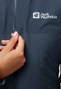 Veste bleu marine avec une texture lisse, dotée d'une fermeture éclair centrale et d'un écusson de logo. Main saisissant le tirette de la fermeture.