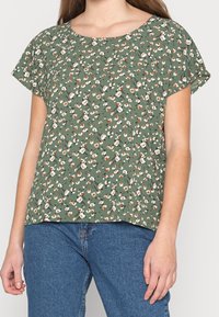 Zielona bluzka w kwiaty z krótkimi rękawami, z zaokrąglonym dekoltem i luźnym fasonem. Wzór zawiera białe i brązowe kwiaty na zielonym tle.