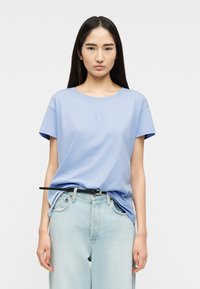 Γυναίκα με μακριά σκούρα μαλλιά, που φοράει ανοιχτό μπλε μπλουζάκι τυλιγμένο μέσα σε ανοιχτό μπλε τζιν με μαύρη ζώνη, στέκεται μπροστά σε απλό φόντο.