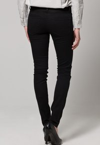 Svarta skinny jeans med en jämn textur, hög midja och bakfickor, parat med svarta högklackade skor, sedd bakifrån.