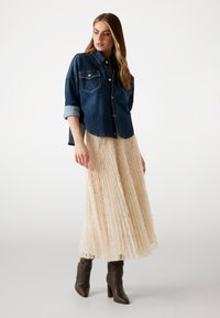Veste en denim avec deux poches poitrine et manches retroussées, associée à une jupe plissée beige, longueur genoux, et des bottes à talons marron.