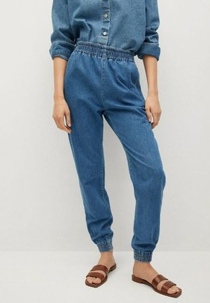 Vrouw draagt blauwe denim joggerbroek met elastische taille, bijpassend denim overhemd met knopen en bruine platte slides.