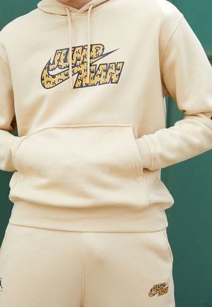 Beżowy komplet bluzy z kapturem i spodni dresowych z logo Nike Air w panterkę na klatce piersiowej i lewym udzie, ręce w przedniej kieszeni bluzy.
