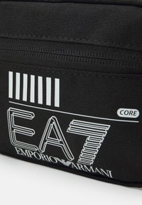 Černá zipová taška s texturovanou látkou. Obsahuje bílé grafické logo „EA7“ a vertikální pruhy. Obsahuje text „EMPORIO ARMANI“. 