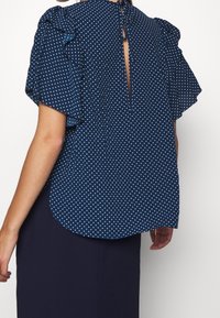 Marinblus med prikkmønster, volangerm, detalj med nøkkelhull bak og avrundet kant. Laget av lett stoff for komfort.