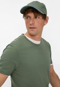 T-shirt di cotone verde con scollo rotondo bianco e maniche corte. Abbinato a un cappellino verde con visiera blu. Nessun altro dettaglio visibile.