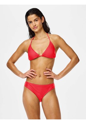 Frau mit nassen Haaren, die ein rotes Bikini-Top in Dreiecksform und passende Unterteile trägt, steht mit den Händen in der Hüfte vor weißem Hintergrund.