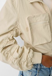 Beige bomber jacket with a zippered chest pocket. Features elasticized cuffs, a textured finish, and ribbed waistband. Lightweight fabric.