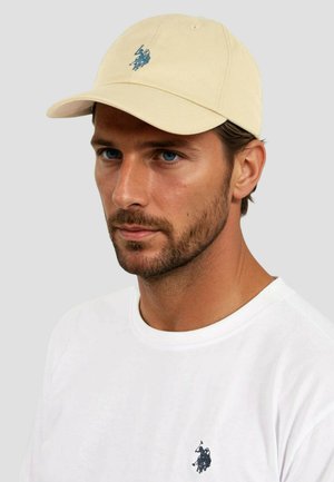 Beige baseball cap with a stitched blue logo at the front, featuring a curved brim and cotton material. Worn with a plain white t-shirt.