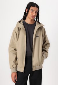 Khaki hooded jacket with a front zipper, elastic cuffs, and a small logo on the left chest, paired with a black shirt and dark pants.