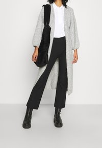 Šedý pletený dlouhý cardigan s balónovými rukávy, spárovaný s černými zvonovými kalhotami a robustními černými kotníkovými botami. Nese texturovanou černou tašku.