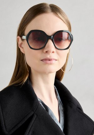 Lunettes de soleil surdimensionnées noires avec des verres teintés en rouge et des montures épaisses. Le modèle porte un manteau noir et de grandes boucles d'oreilles en or.