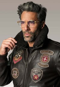 Homme aux cheveux poivre et sel et portant des lunettes, ajustant le col d'une veste en cuir noir ornée de patchs et de badges militaires.