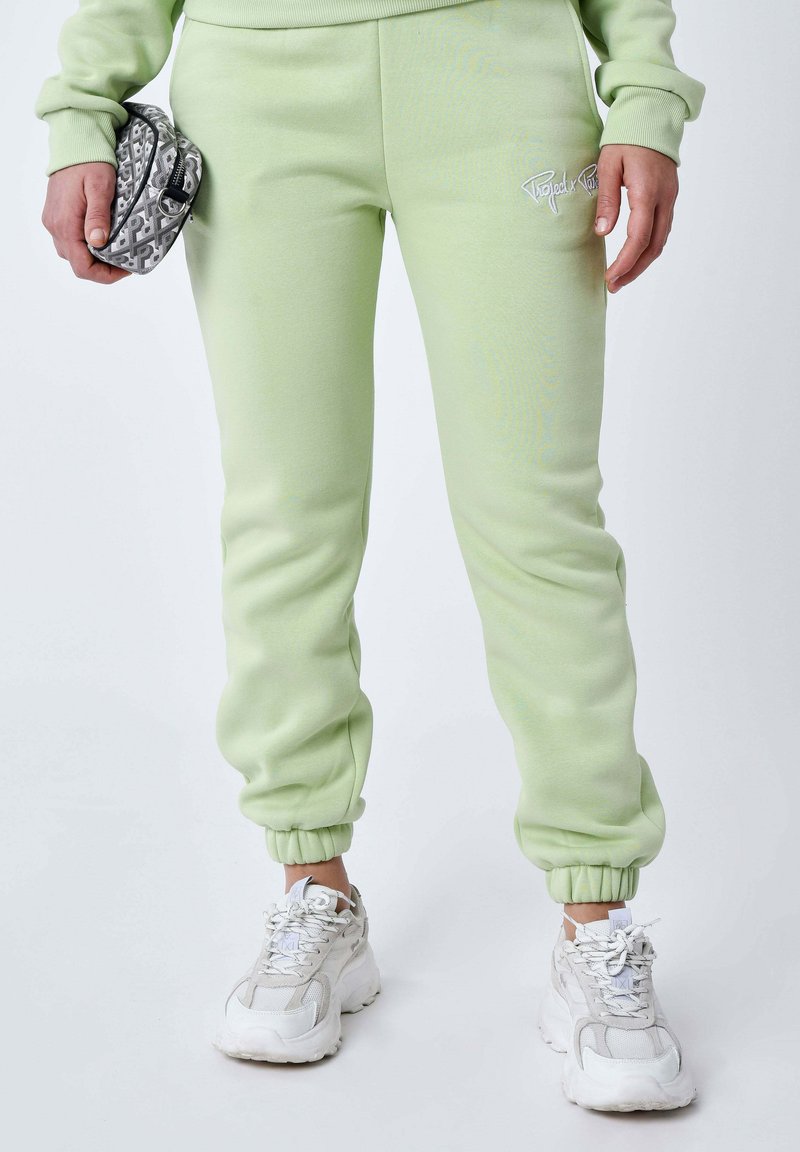 Pantalones deportivos verde claro con puños acanalados, que presentan un logo bordado. Se sostiene una bolsa gris con estampado, complementando unas zapatillas blancas de suela gruesa.
