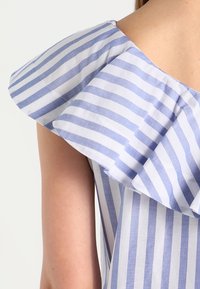Blusa a righe blu e bianche con un design a spalle arricciate. Realizzata in tessuto leggero con una superficie liscia.