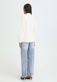 Blazer blanc à manches longues, présentant une coupe ajustée. Associé à un jean bleu clair taille haute à jambes larges et des mules marron à talons.