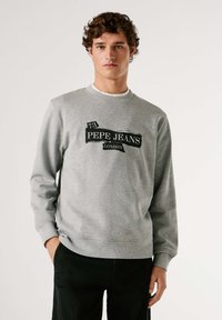 Felpa grigia con polsini e orlo a coste, con logo grafico nero "PEPE JEANS LONDON" al centro. Indossata sopra una camicia bianca.