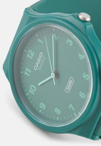 Mátově zelené quartz hodinky Casio s kulatým ciferníkem, průhledným sklem, jednoduchými číslicovými ukazateli a odpovídajícím měkkým silikonovým páskem. Indikována vodotěsnost.