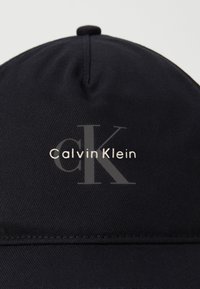 Casquette de baseball noire en tissu lisse avec un logo en relief en texte argenté : "Calvin Klein" et un grand 'CK' en gris.