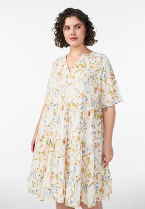 Žena s kučeravými vlasmi nosí voľné, biele šaty po kolená s krátkymi rukávmi a viacfarebným kvetinovým vzorom.