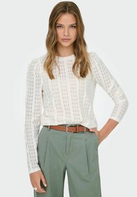 White long-sleeve top made of lace-like fabric with vertical patterns, paired with grey trousers and a brown belt. Textured, lightweight design.