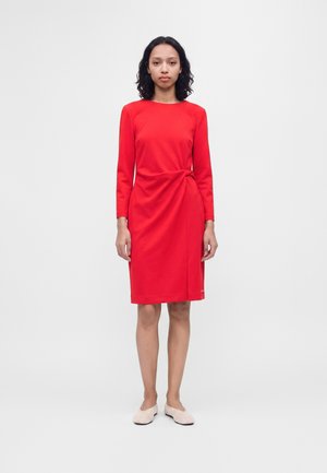 Femme aux cheveux foncés mi-longs, portant une robe rouge à manches longues jusqu'aux genoux et des chaussures plates blanches, debout devant un fond uni.
