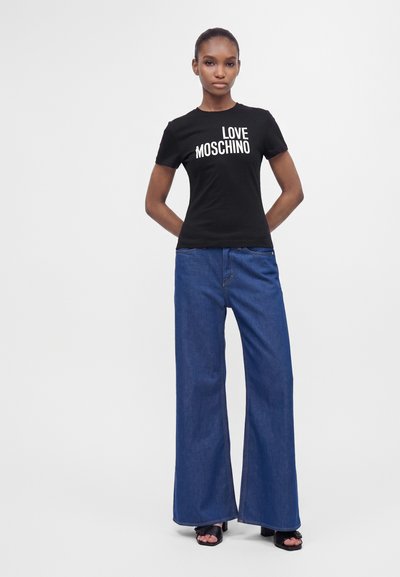 Жена, облечена в черна тениска Love Moschino, широки сини дънки и черни обувки с отворени пръсти, стои с ръце зад гърба на бял фон.