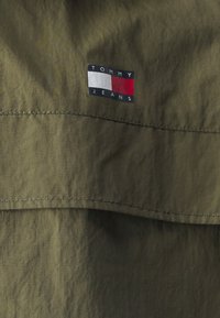 Olivgrön tyg med en slät textur har en rektangulär logotyp-patch med "TOMMY JEANS" och en tri-färgad design i röd, vit och blå.