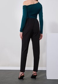 Teal off-shoulder top with long sleeves and gathered detail, paired with black fitted trousers and black heeled shoes. Minimalist setting.