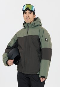 Grüne und schwarze isolierte Jacke mit Kapuze, Reißverschlusstaschen und Markenlogo. Getragen mit einem schwarzen Helm und verspiegelt Sonnenbrille.