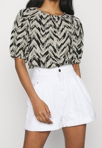 Blusa a zigzag nera e beige con maniche a palloncino, abbinata a pantaloni corti bianchi a vita alta con piega all'orlo e bottone frontale.