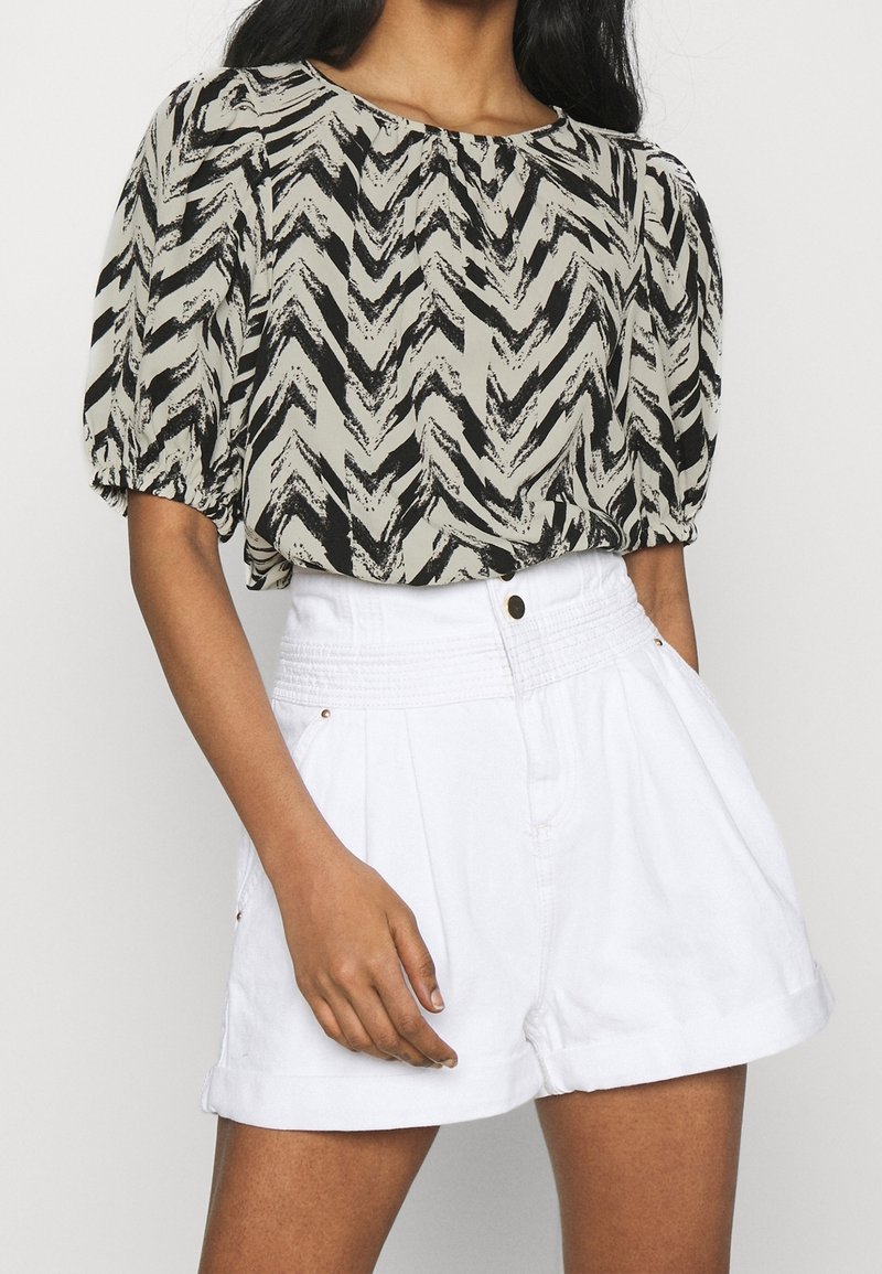Blusa a zigzag nera e beige con maniche a palloncino, abbinata a pantaloni corti bianchi a vita alta con piega all'orlo e bottone frontale.