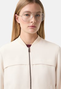 Jasnokremowa bomberka z zamkiem z przodu, prążkowanym kołnierzem i subtelnymi detalami panelowymi. Modelka nosi przezroczyste, oversize'owe okulary.