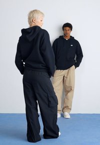 Deux mannequins portent des vêtements de sport coordonnés. L'un est en sweat à capuche noir et en pantalon cargo noir ; l'autre est en sweat à capuche noir et en pantalon beige.