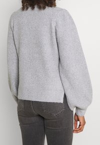 Person som bär en ljusgrå stickad tröja med något puffiga ärmar och mörkgrå åtsittande jeans, sett bakifrån.