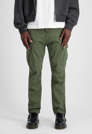 Cargo pants in olive green cotton, slim fit, with multiple side pockets. Paired with black lace-up boots, featuring yellow stitching accents.